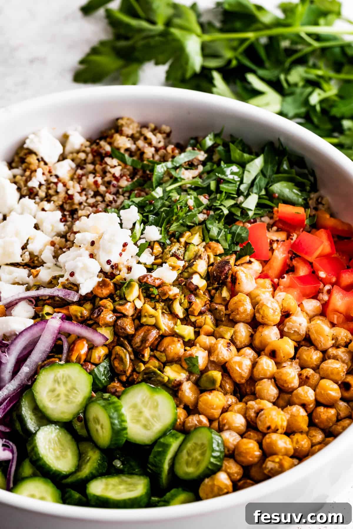 A large bowl with sliced cucumbers, sliced red onions, chickpeas, diced red bell peppers, herbs, tri-colored quinoa, and crumbles of feta cheese.