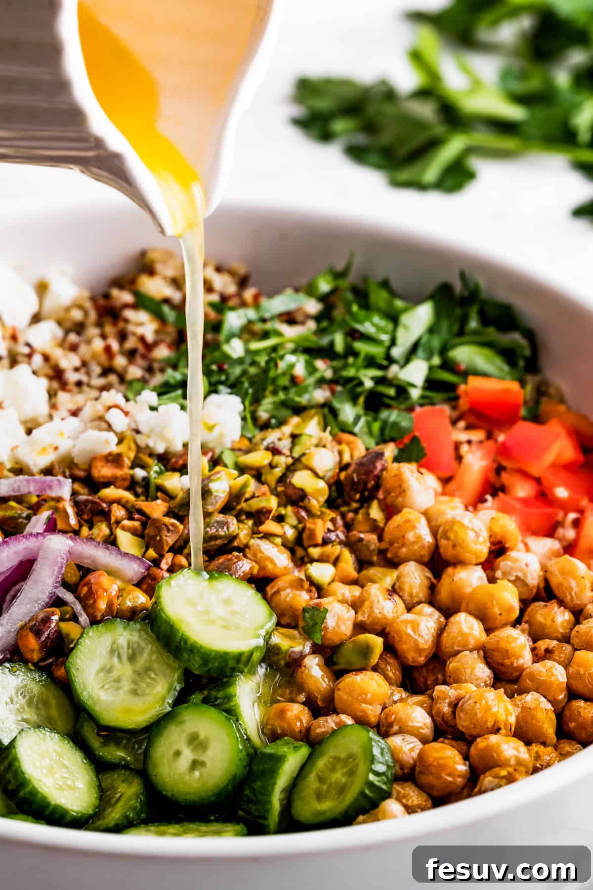 Pouring salad dressing over chickpeas, sliced cucumbers, red onions, peppers, herbs, and feta.