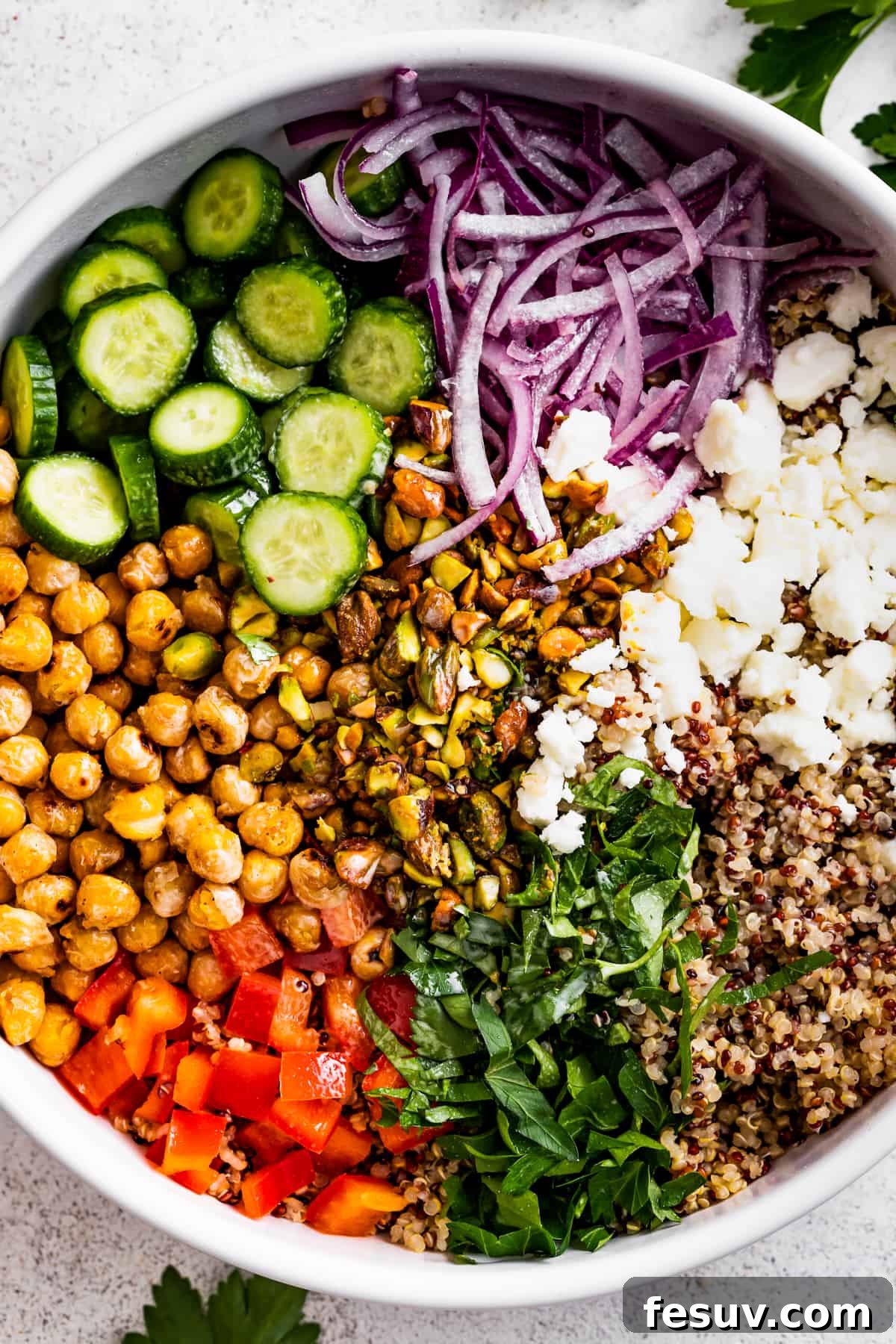 A large bowl with sliced cucumbers, sliced red onions, chickpeas, diced red bell peppers, herbs, tri-colored quinoa, and crumbles of feta cheese.