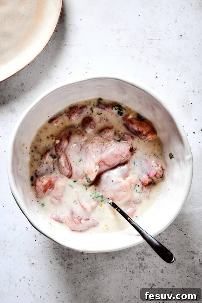Coating chicken thighs in the flavorful yogurt marinade. Boneless, skinless chicken thighs being added to a rich, herbaceous yogurt marinade in a bowl, ensuring full coverage before refrigeration.