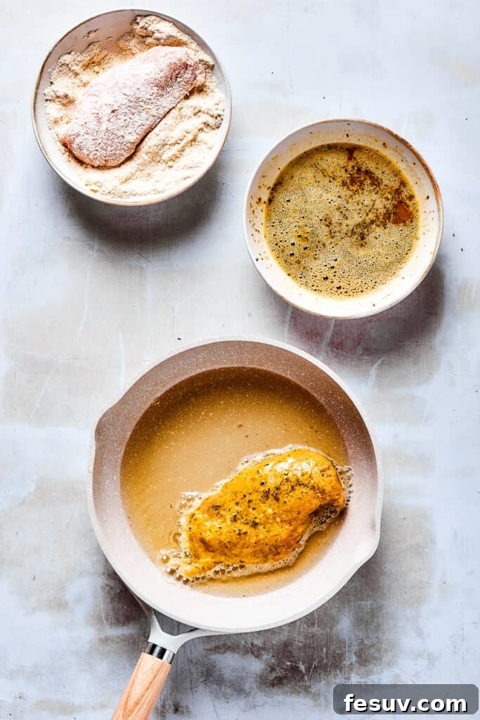 Starting our Lemon Chicken: Crispy, parmesan-crusted chicken with real lemon butter sauce. A raw chicken breast is coated in a dish of dry breading, adjacent to a bowl of wet egg mixture, with a skillet on the stove containing a cooking breaded chicken breast.