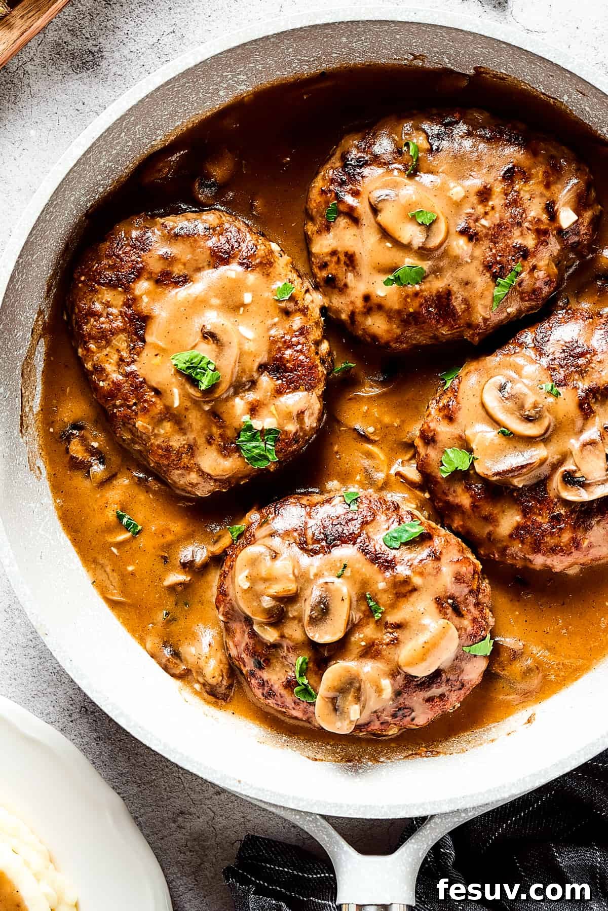 Four perfectly cooked Salisbury steaks drenched in a rich mushroom gravy, served in a cast-iron skillet. The dish is garnished with fresh herbs, alongside vibrant vegetables and a rustic cloth napkin, evoking a cozy, homestyle meal.