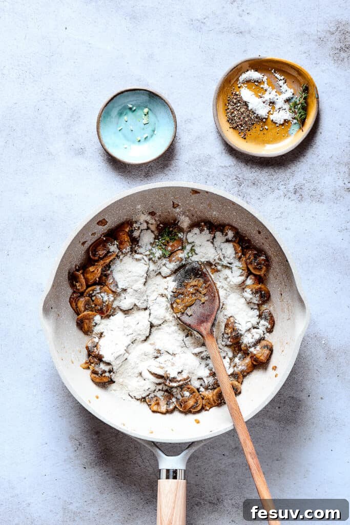 All-purpose flour is evenly sprinkled over the sautéed mushroom and onion mixture in a skillet, forming the base for a rich roux to thicken the gravy.