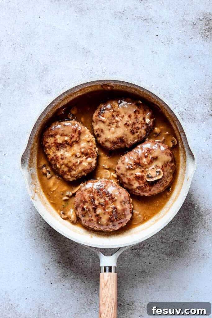 The finished Salisbury steak dish in a skillet, with golden-brown patties nestled in a thick, savory mushroom gravy, garnished with fresh herbs, ready to be served.