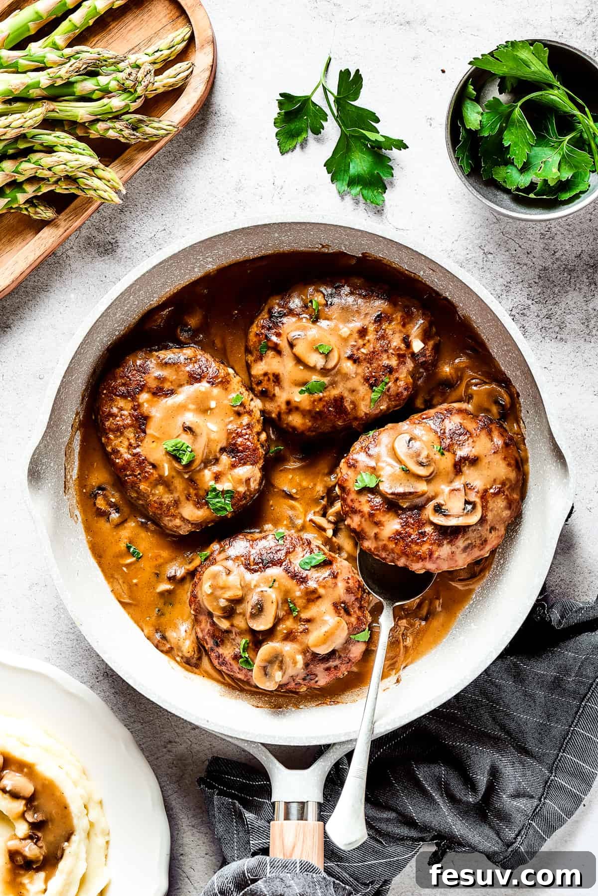 Four golden-brown Salisbury steaks, generously covered in a rich mushroom gravy, presented beautifully in a rustic cast-iron skillet. The dish is artfully arranged with fresh rosemary sprigs, colorful vegetables like carrots and green beans, and a neatly folded linen napkin for an inviting, homestyle meal.