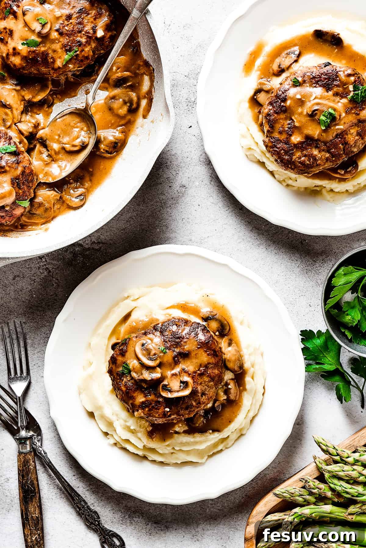 A cast-iron skillet holds several Salisbury steaks submerged in gravy, flanked by two dinner plates each with a steak and mashed potatoes, highlighting a complete and satisfying meal presentation.