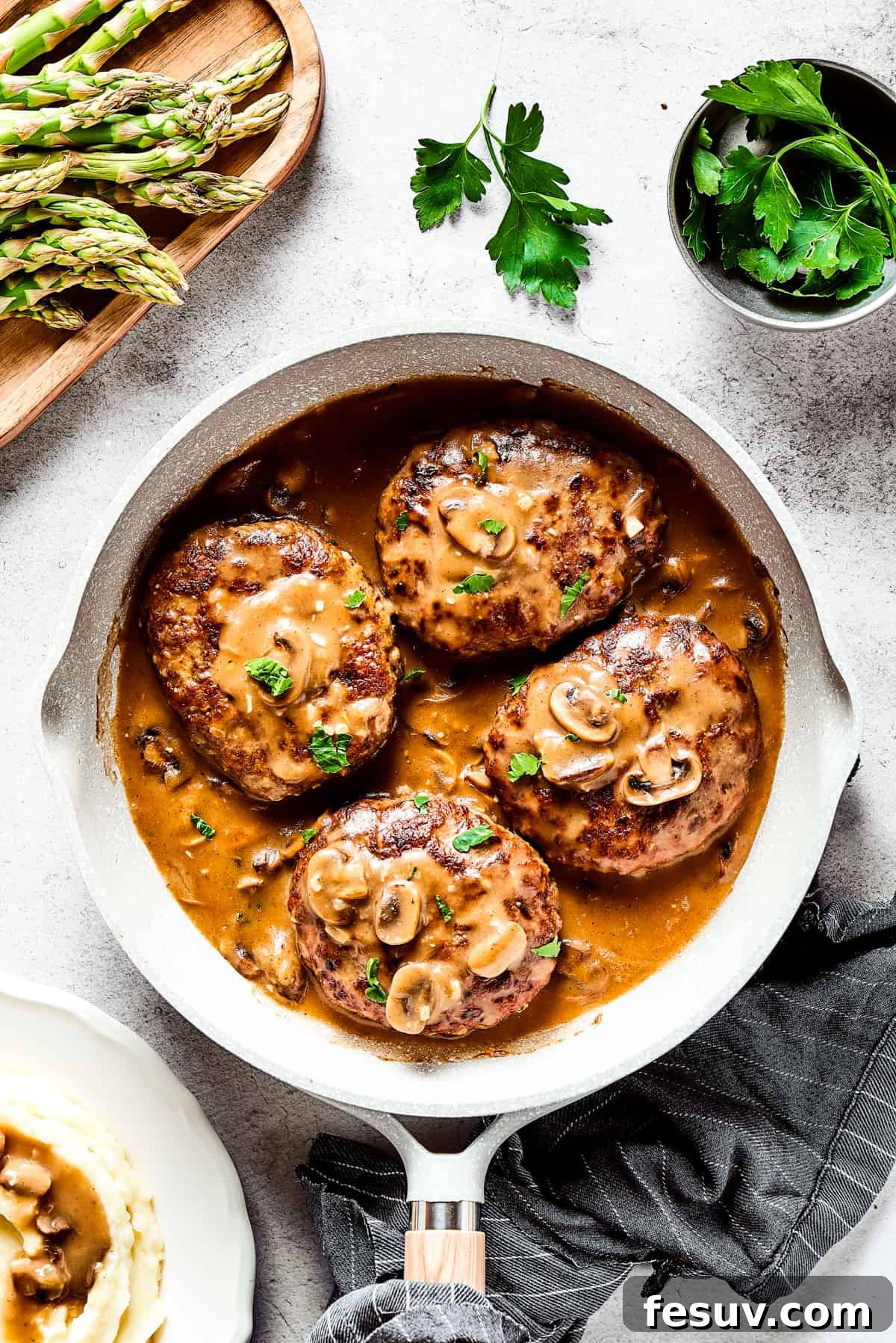 A close-up shot of four golden-brown Salisbury steaks simmering in a rich, dark mushroom gravy within a cast-iron skillet, garnished with fresh thyme. The rustic presentation highlights the hearty, homemade nature of the dish.