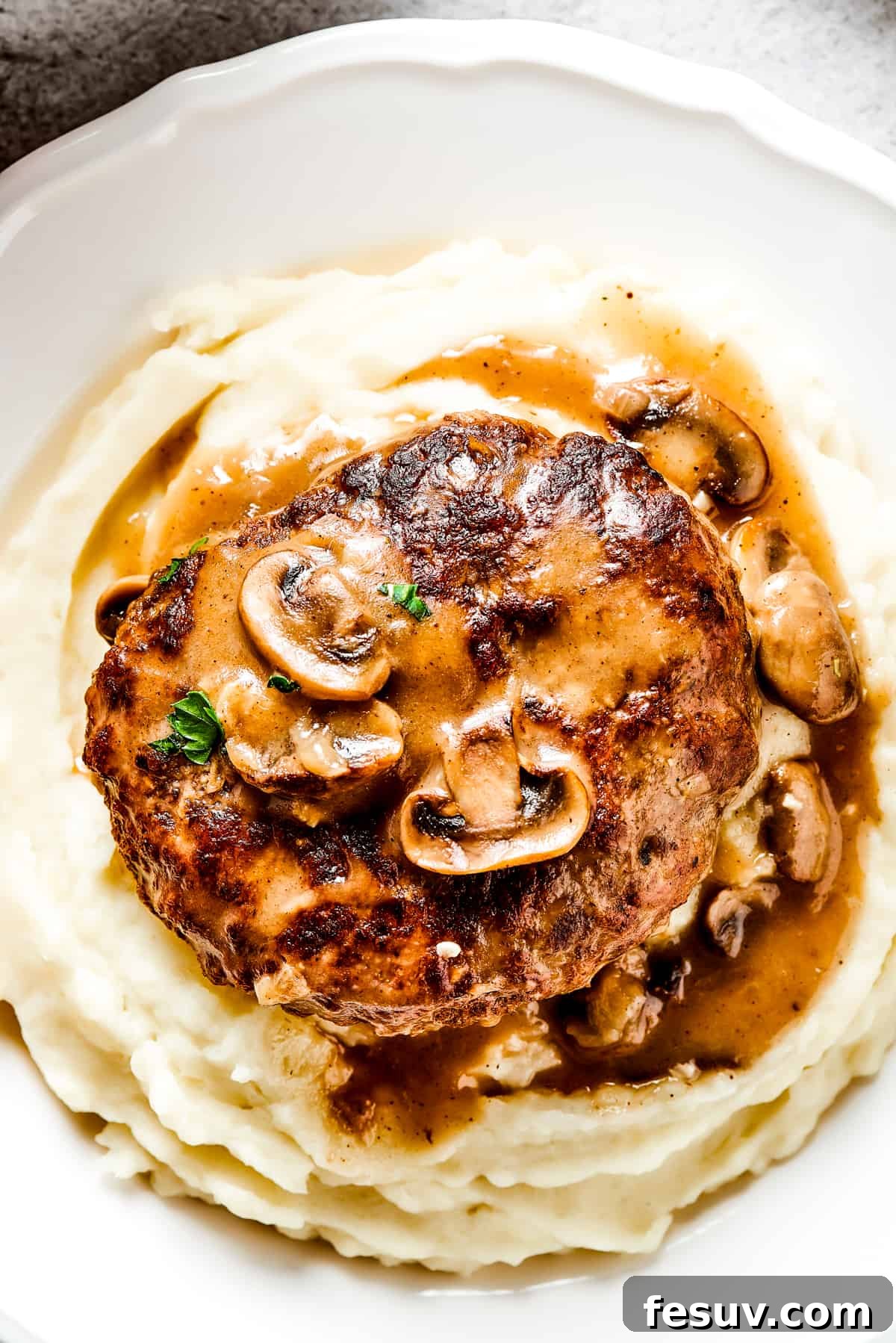 A perfectly plated Salisbury steak, glistening with rich mushroom gravy, served atop a generous bed of creamy mashed potatoes. Fresh green herbs garnish the dish, adding a touch of color and freshness.