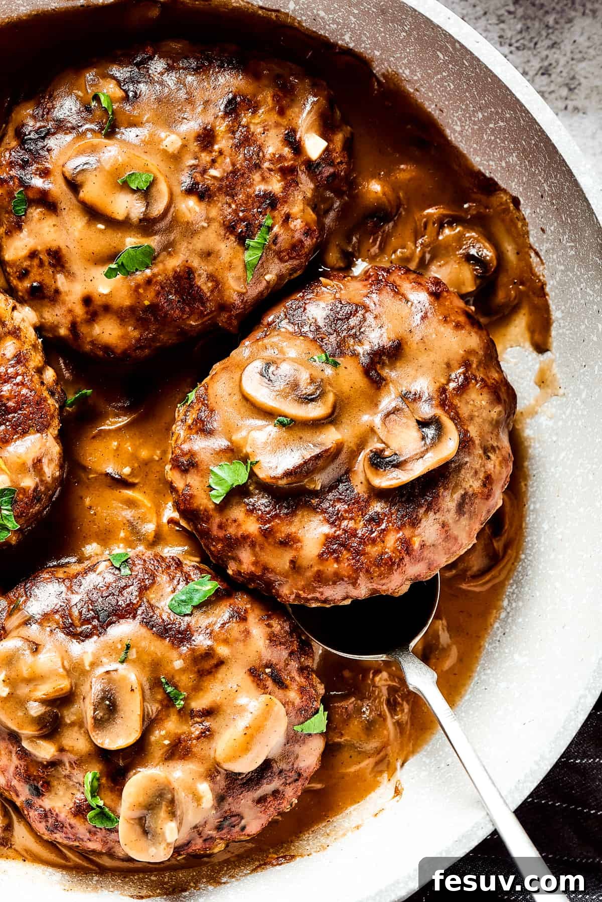 A close-up shot of a cast-iron skillet filled with four Salisbury steaks swimming in a bubbling, rich mushroom and onion gravy, garnished with fresh green herbs. The rustic presentation emphasizes the hearty, homemade quality of the dish.