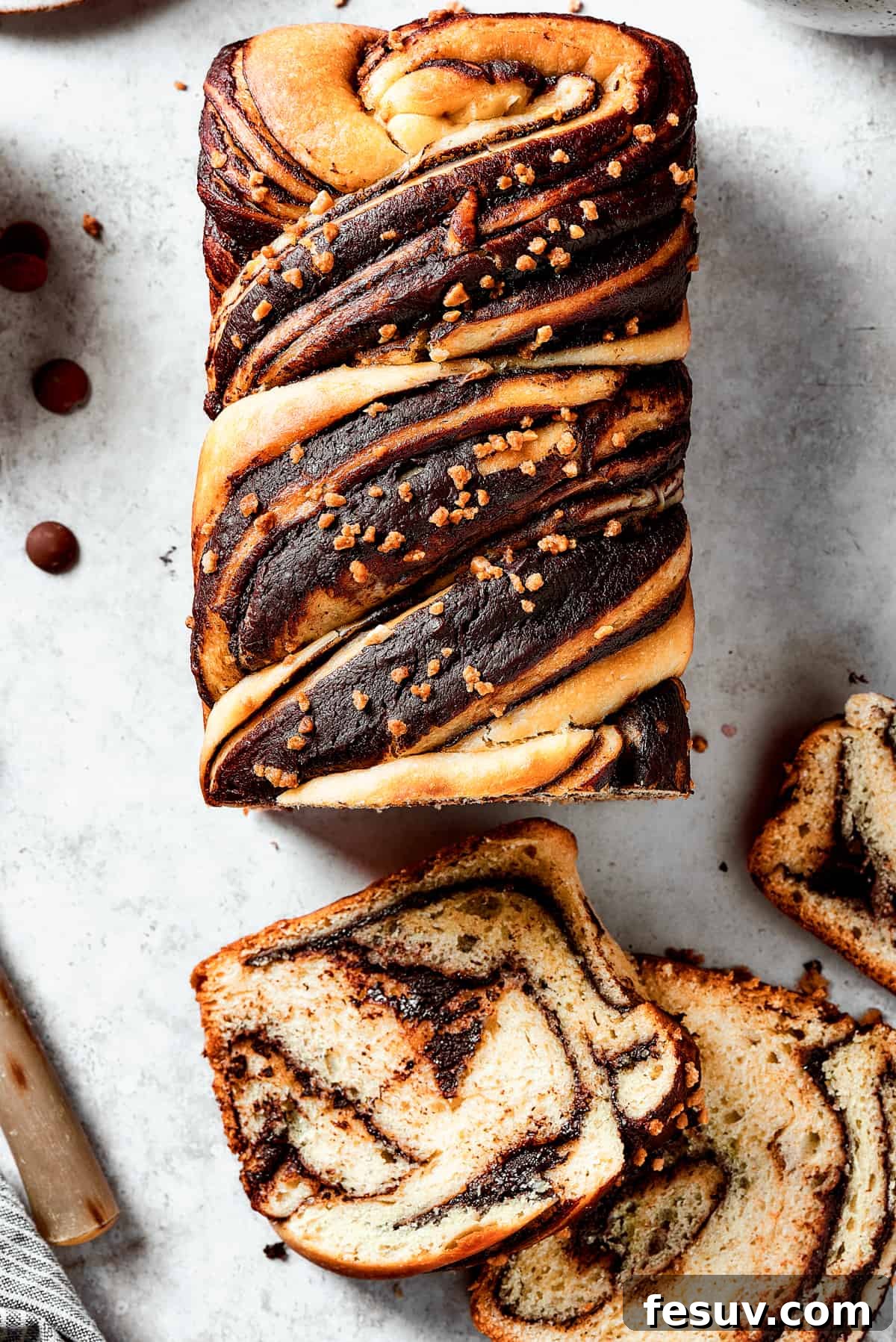 A loaf of chocolate babka that has been partially sliced. The slices are laid out on the work surface, along with a bread knife and a dish of chocolate chips.