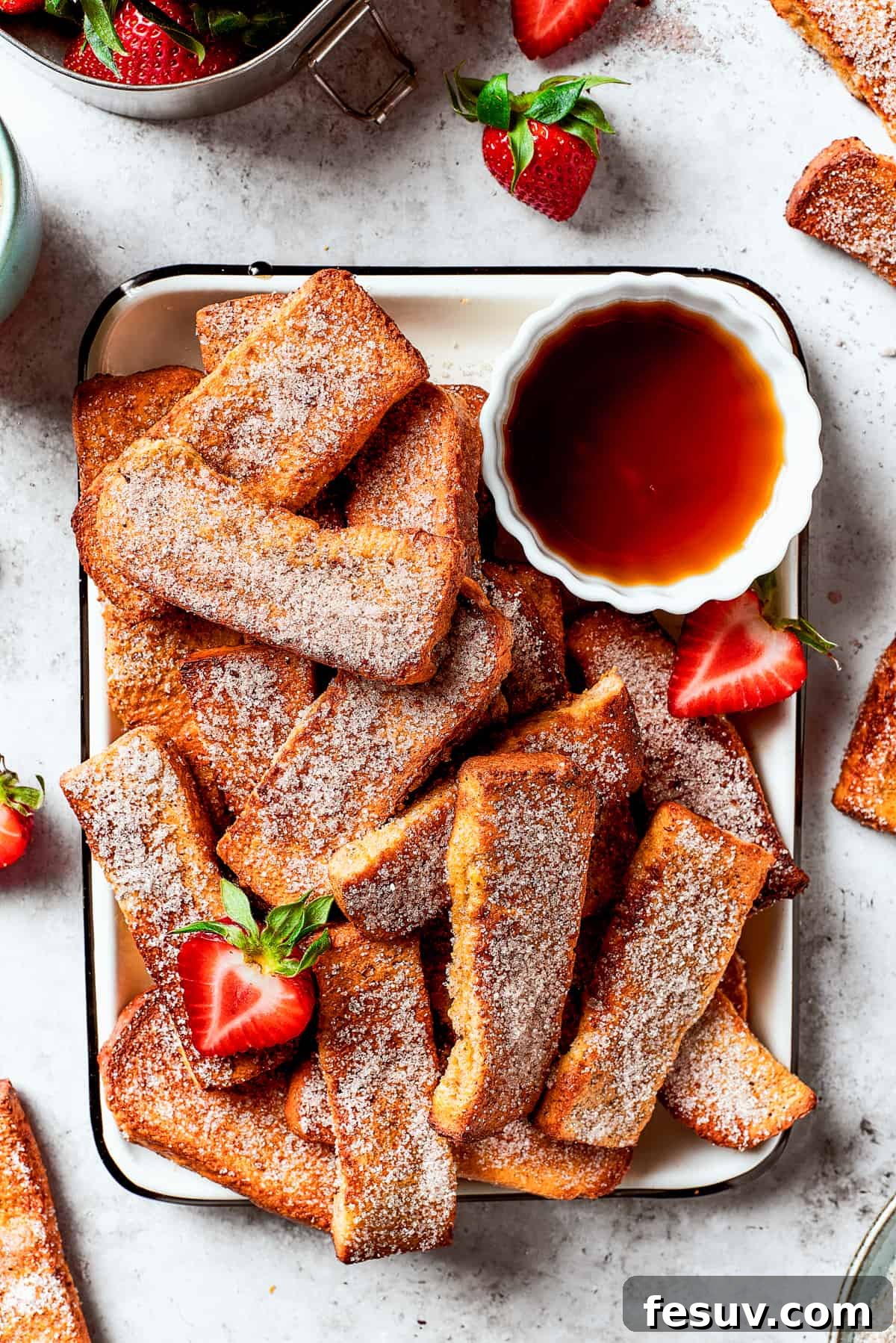 Air fryer French toast sticks on an enamel tray with a cup of maple syrup and fresh strawberries.