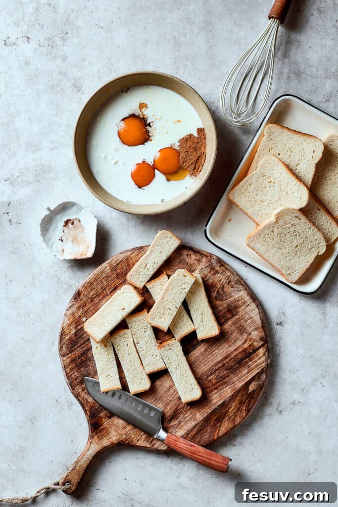 A bowl of eggs, milk, and other custard ingredients next to a tray of sliced bread, and a cutting board of additional bread cut into sticks.
