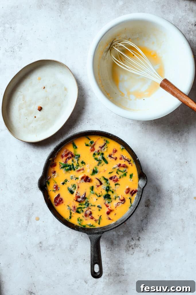 Spinach, Bacon, and Potato Frittata is a healthy, farmhouse-style dinner that's easy to make, and loaded with rich flavor. Perfect easy meal! Pouring egg scramble over bacon, onion, spinach, and potatoes in a cast-iron pan.