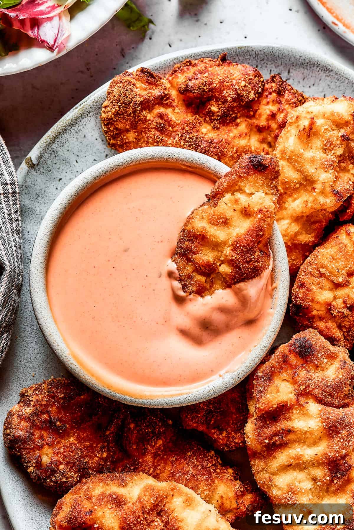 A platter of chicken strips with one piece dipped in a bowl of sauce.