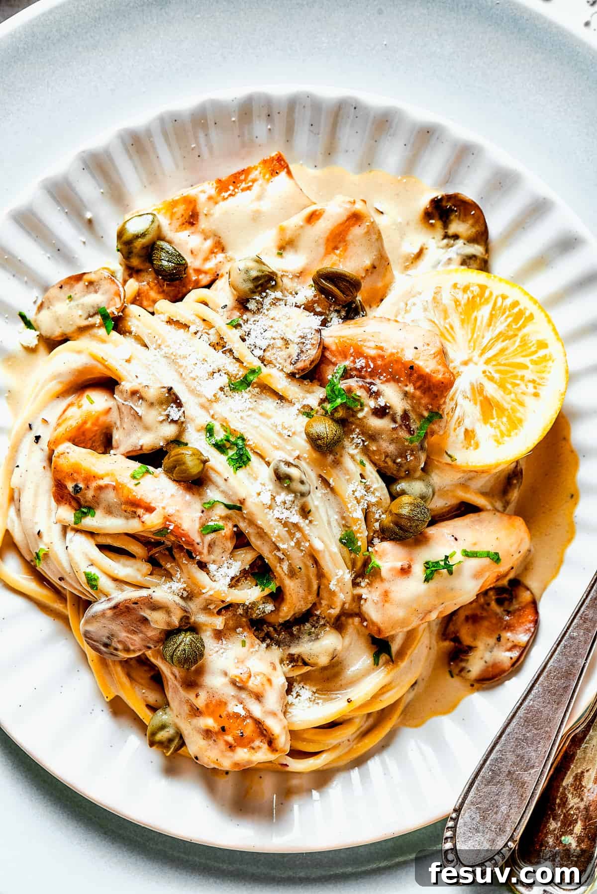 Experience the decadent flavors of Cheesecake Factory Chicken Piccata with this perfect homemade copycat recipe. Chicken piccata with pasta, mushrooms, and lemon slices in a creamy caper sauce, served on a white dinner plate.