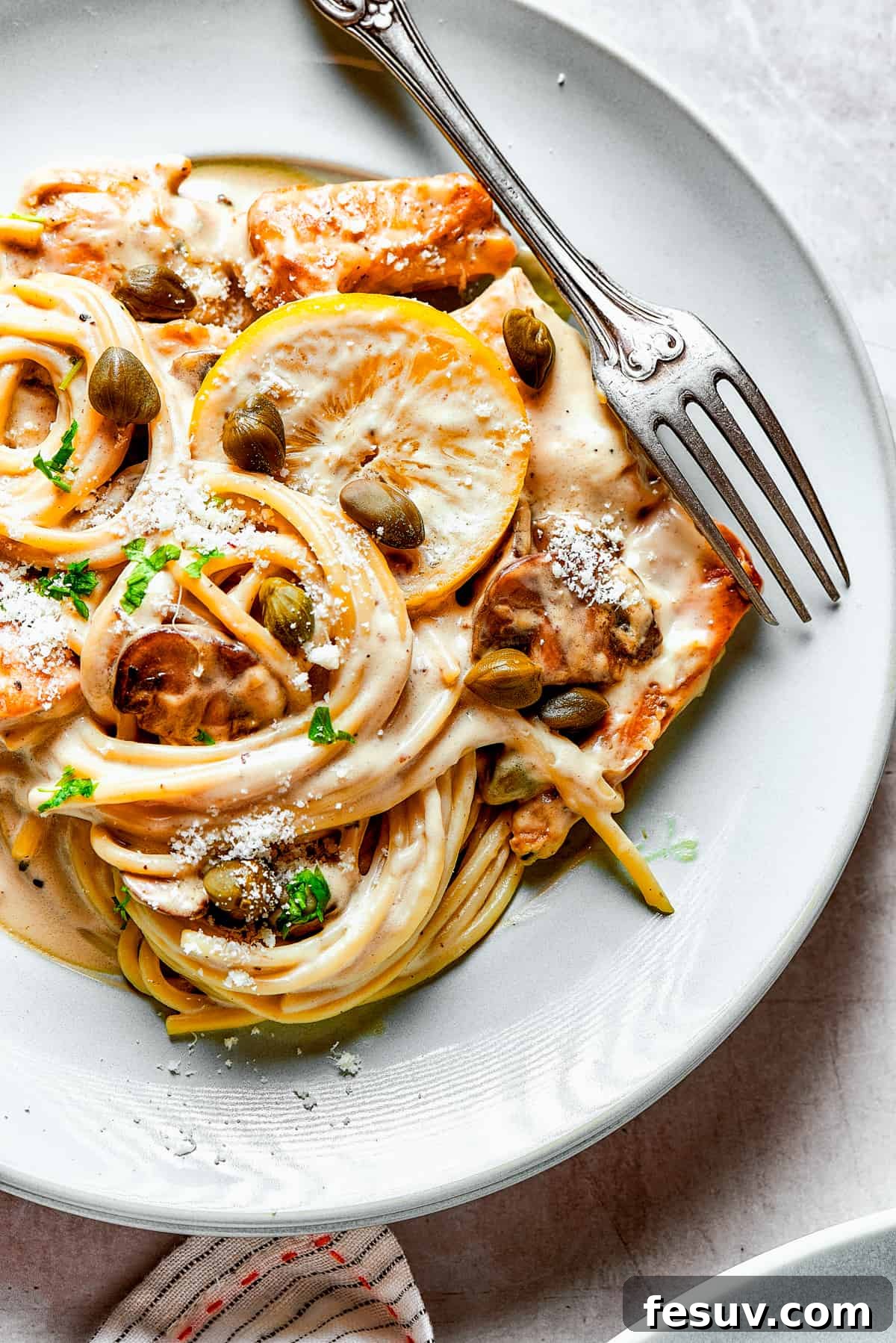 A tempting overhead view of the creamy chicken pasta. Overhead view of a generous serving of Chicken Piccata pasta on a white dinner plate with a fork, ready to be eaten.