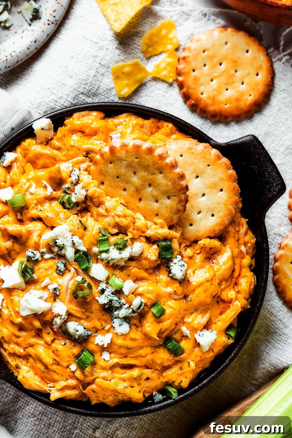 Overhead shot of a serving of Crockpot Buffalo Chicken Dip with crunchy crackers, fresh blue cheese crumbles, and vibrant green onions as garnish.