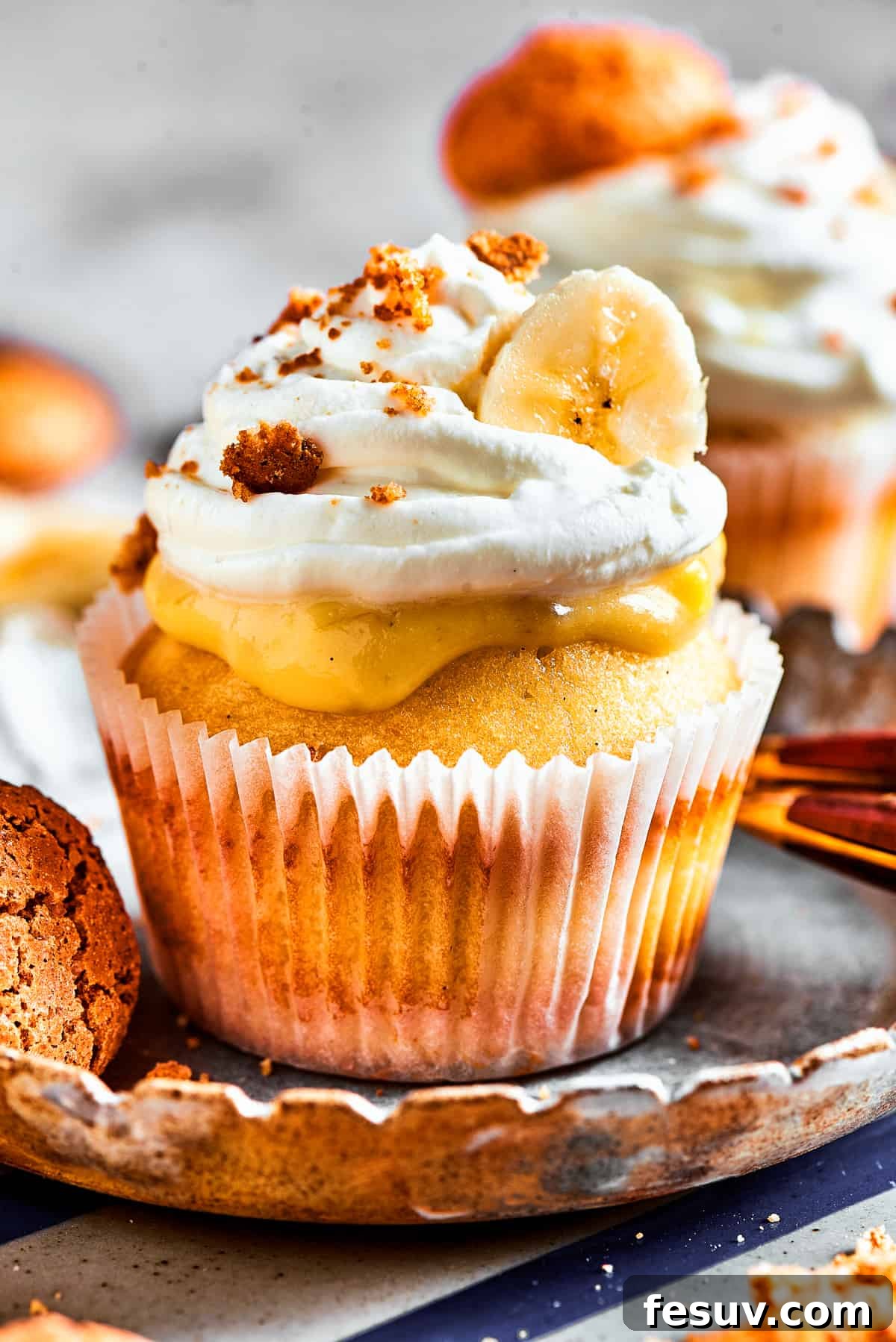 Banana Pudding Cupcakes: A sweet surprise in a small package! One banana pudding cupcake, beautifully presented on a dessert plate with more cupcakes in the blurred background. It features a fluffy cake base, creamy banana pudding filling, and toppings of whipped cream, a fresh banana slice, and a vanilla wafer.