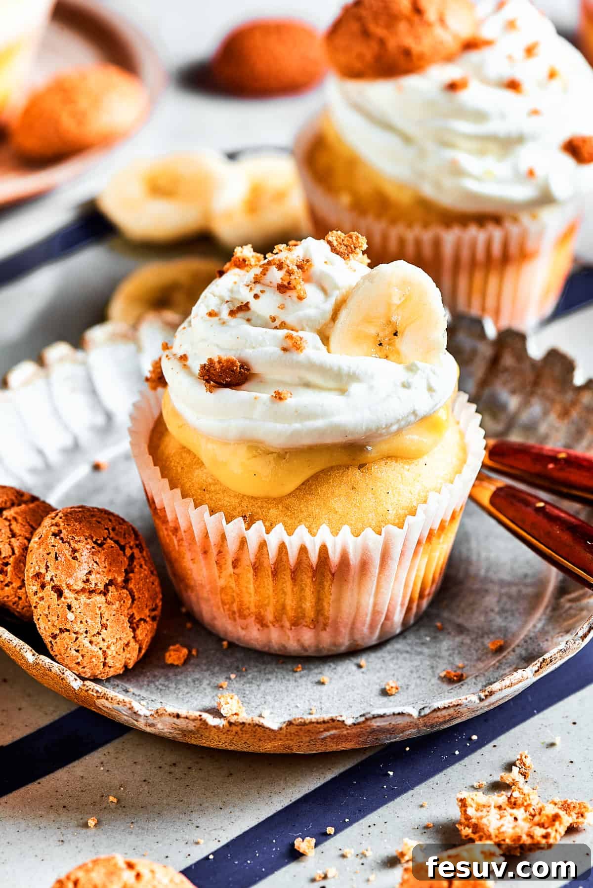 Fluffy cupcakes filled with creamy pudding and topped with perfect banana pudding garnishes. One banana pudding cupcake on a dessert plate, with a slightly different angle from the first, showing the inviting texture. More cupcakes are blurred in the background.