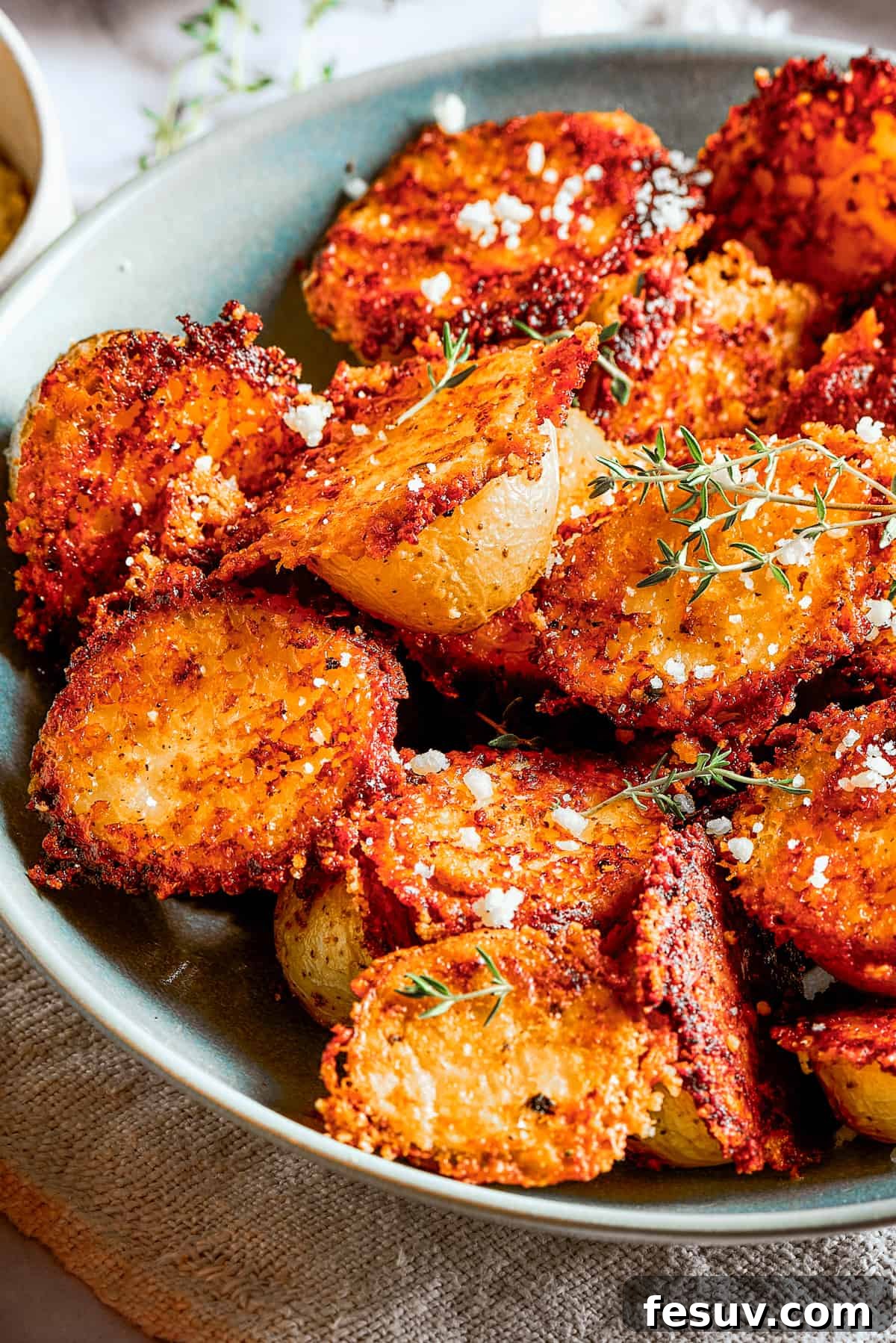 These tender potatoes with a crunchy, cheesy crust are the easiest, tastiest side dish you'll love! Cheesy roasted potatoes in a bowl next to cups of dipping sauce, a cloth napkin, and a serving on a plate.