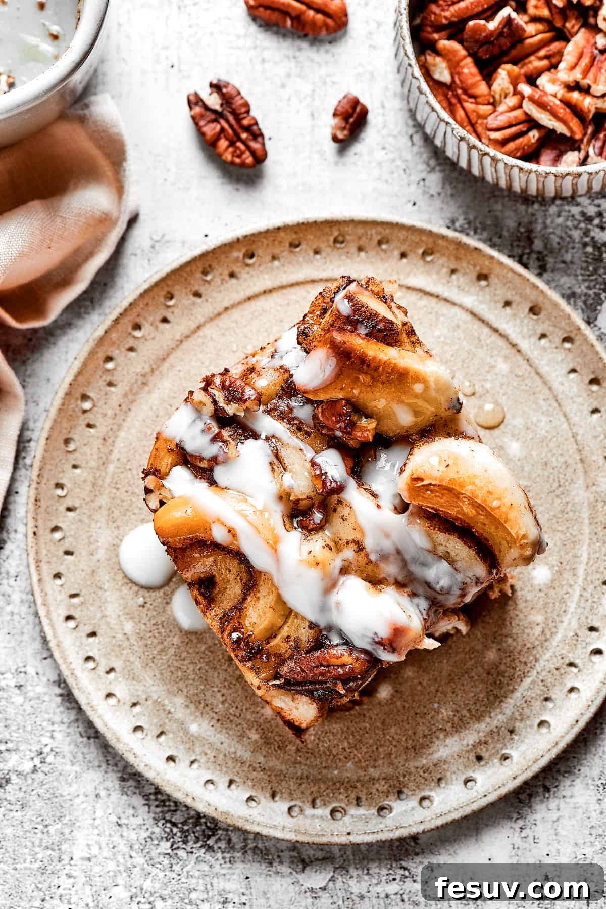 These easy and celebratory cinnamon roll casseroles are rich, creamy, and buttery, and packed with sticky and sweet maple syrup and pecans. An inviting overhead view of a single slice of cinnamon roll casserole elegantly placed on a white plate, showcasing its golden crust, soft interior, and drizzled icing. A small bowl of fresh pecans sits beside it.