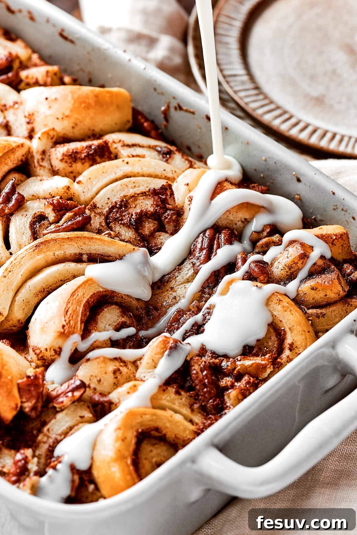 These easy and celebratory cinnamon roll casseroles are rich, creamy, and buttery, and packed with sticky and sweet maple syrup and pecans. A close-up of a freshly baked cinnamon roll casserole with a bowl of white icing being gently drizzled over its warm, golden surface, creating beautiful, enticing patterns.