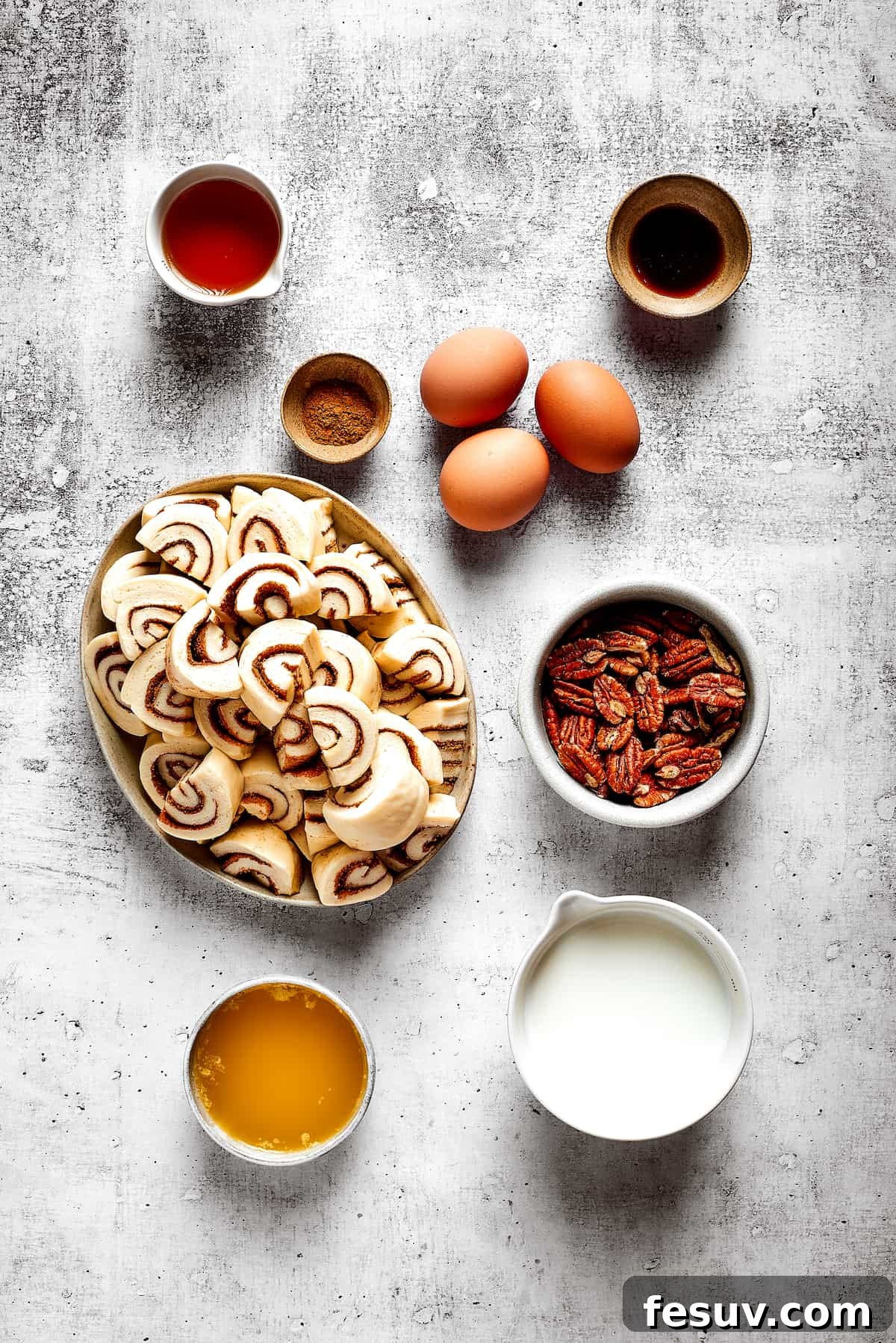 These easy and celebratory cinnamon roll casseroles are rich, creamy, and buttery, and packed with sticky and sweet maple syrup and pecans. An inviting overhead spread of all the fresh ingredients needed for a delicious cinnamon roll casserole. Featured are a plate of quartered cinnamon roll pieces, a bowl of creamy milk, rich maple syrup, crunchy pecans, three eggs, ground cinnamon, pure vanilla extract, and a bowl of melted butter.