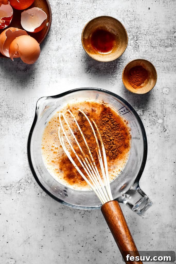 These easy and celebratory cinnamon roll casseroles are rich, creamy, and buttery, and packed with sticky and sweet maple syrup and pecans. A clear glass mixing bowl containing a creamy mixture of eggs and milk, with a dusting of cinnamon on top, ready to be whisked. A whisk rests inside the bowl, with other empty bowls and eggshells in the background.