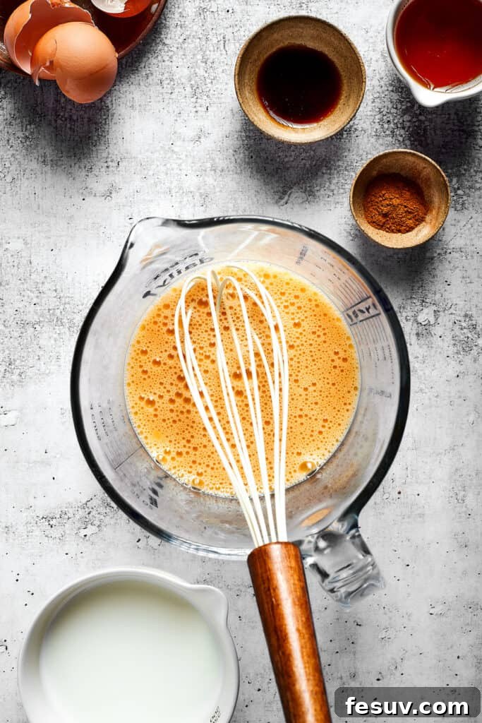These easy and celebratory cinnamon roll casseroles are rich, creamy, and buttery, and packed with sticky and sweet maple syrup and pecans. A glass mixing bowl holds a perfectly whisked, pale yellow egg and milk mixture, ready for the next step. A whisk is still in the bowl, next to other kitchen bowls, indicating an ongoing cooking process.