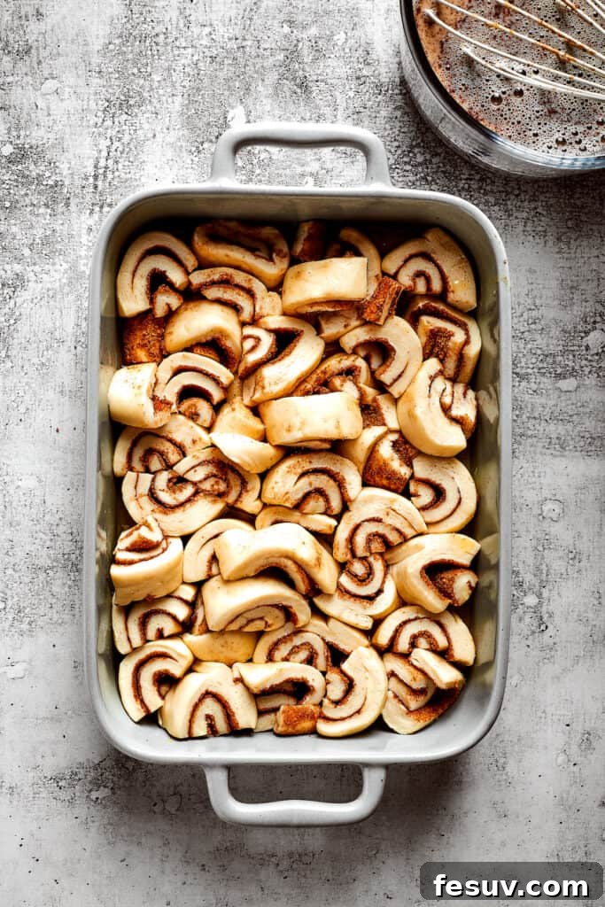 These easy and celebratory cinnamon roll casseroles are rich, creamy, and buttery, and packed with sticky and sweet maple syrup and pecans. An overhead shot of unbaked cinnamon roll pieces arranged neatly in a baking dish, glistening with the recently poured egg and milk mixture, ready for the oven.