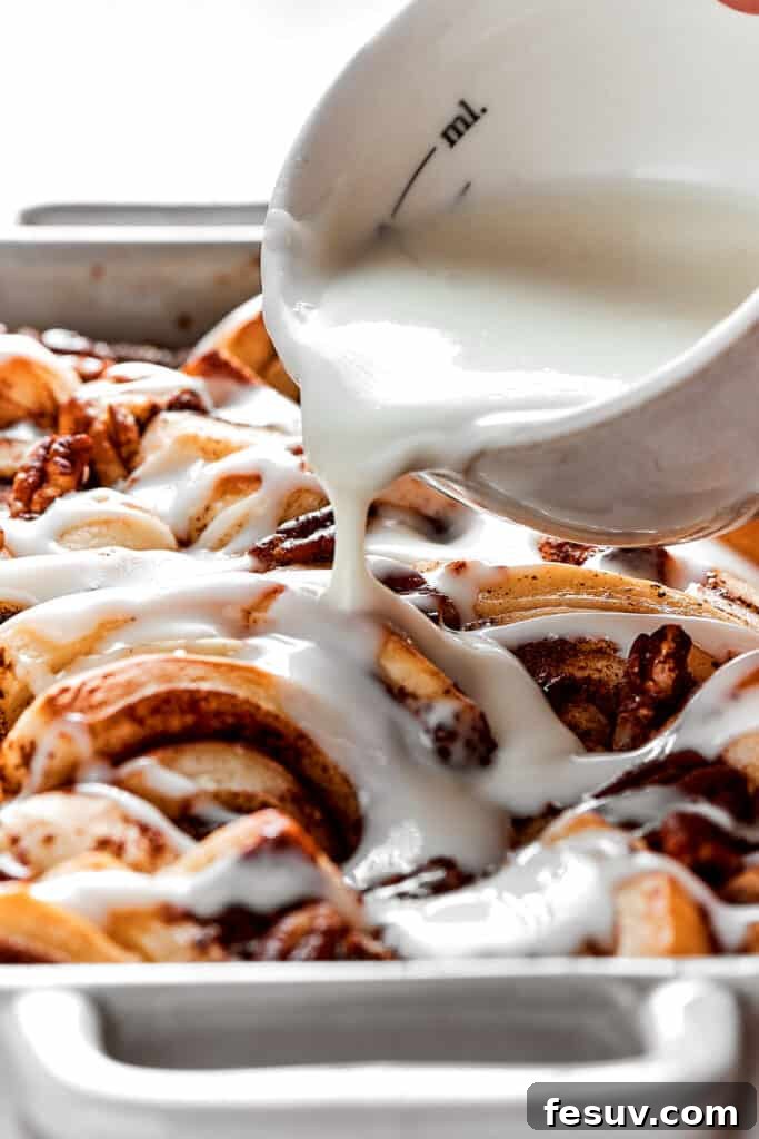 These easy and celebratory cinnamon roll casseroles are rich, creamy, and buttery, and packed with sticky and sweet maple syrup and pecans. A close-up shot capturing the moment a bowl of luscious white icing is being expertly drizzled over the warm, golden-brown cinnamon roll casserole, creating enticing swirls.