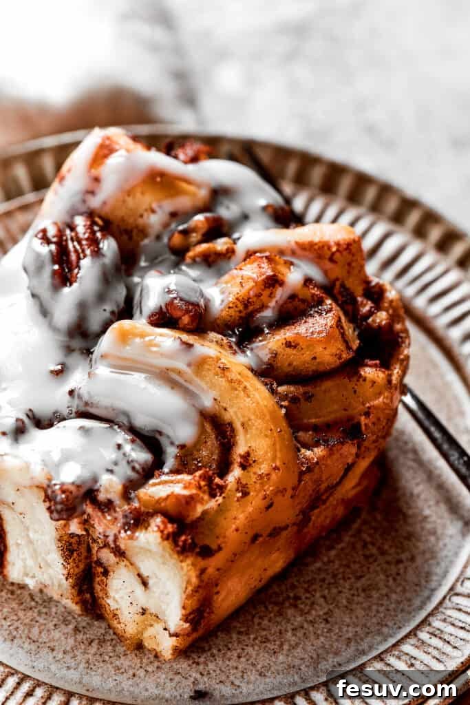 These easy and celebratory cinnamon roll casseroles are rich, creamy, and buttery, and packed with sticky and sweet maple syrup and pecans. A tempting close-up of a square of warm cinnamon roll casserole on a pristine white plate, with a fork poised to capture a bite. The casserole looks perfectly moist and gooey, highlighting its irresistible appeal.