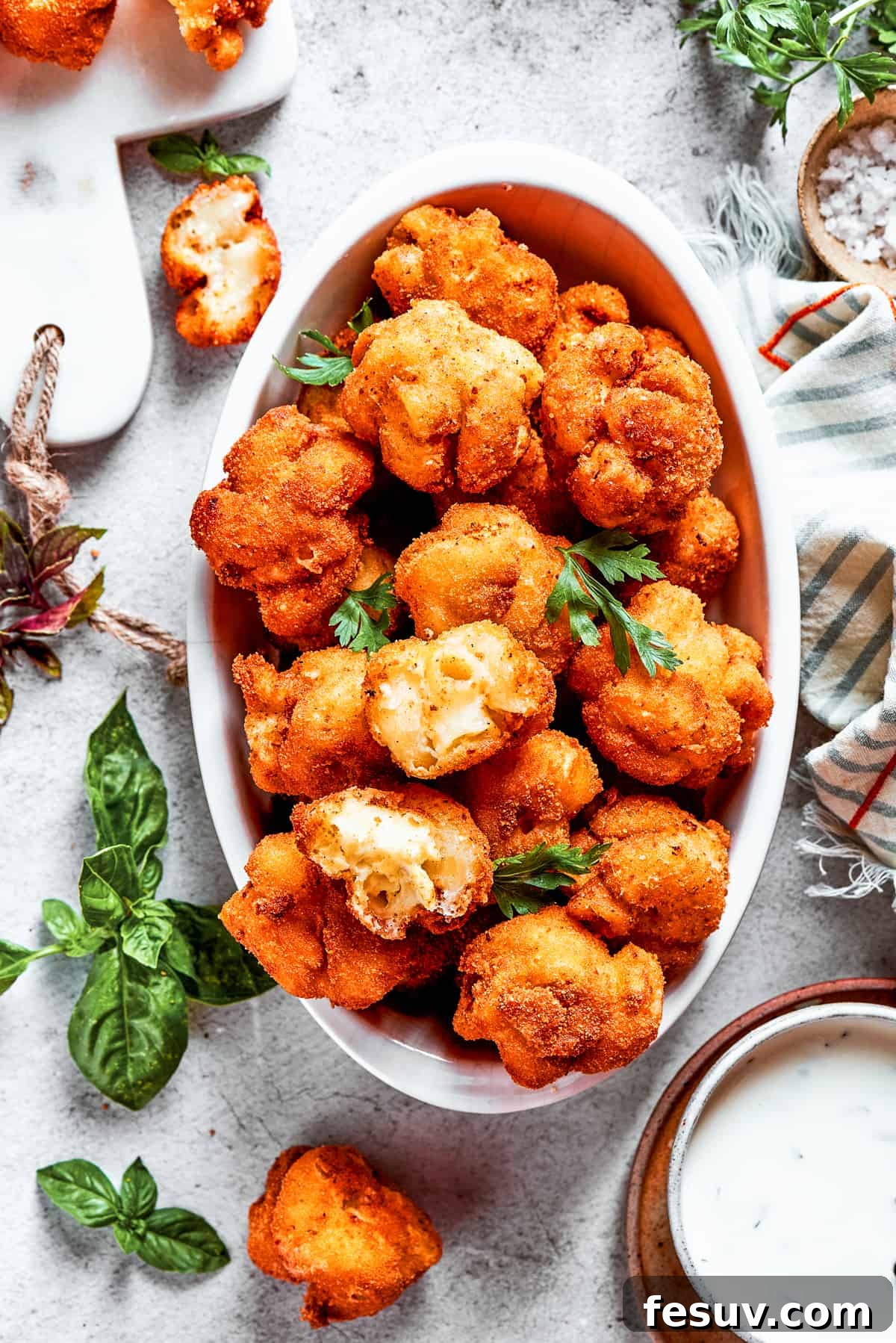 An overhead view of a serving bowl filled with golden-brown mac and cheese bites, artfully arranged next to more bites and garnished with fresh green herbs.