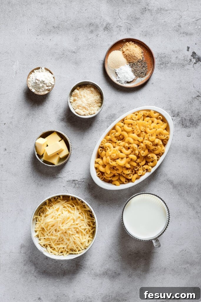 An overhead shot displaying all the ingredients for mac and cheese: a bowl of uncooked elbow macaroni, a bowl of finely shredded mozzarella cheese, a bowl of freshly grated parmesan, a glass of whole milk, a bowl of all-purpose flour, a bowl of unsalted butter, and a small bowl containing salt, pepper, garlic powder, and onion powder.