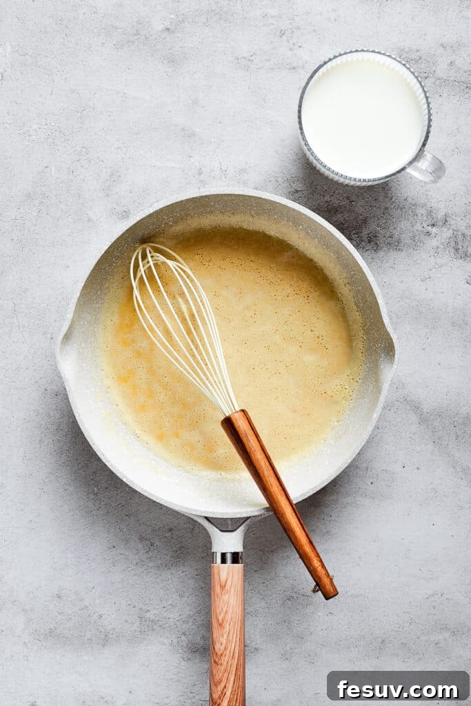 A saucepan on a stovetop, containing a pale, smooth roux, with a metal whisk resting inside the pan.