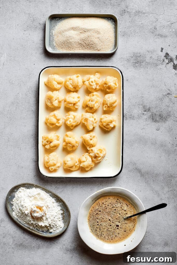 An overhead view of a baking tray filled with perfectly rolled mac and cheese balls, alongside three bowls for the breading station: one with flour, one with breadcrumbs, and one with whisked eggs seasoned with Italian seasoning.