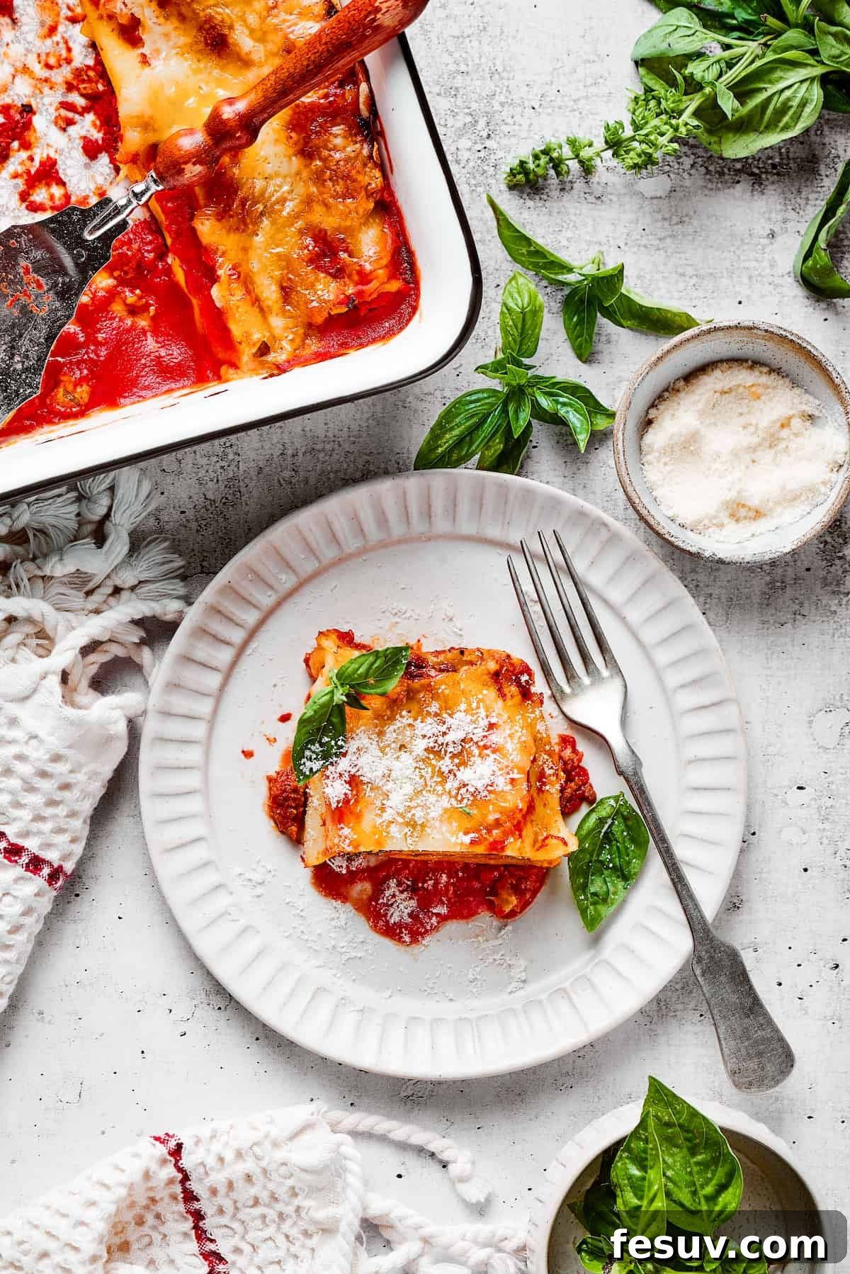 Easy lasagna roll ups have a beefy and herbaceous tomato sauce, a creamy cheese filling, tender pasta, and tons of melted cheese. Overhead view of a lasagna roll up on a plate with a fork, next to a baking dish full of them and some basil leaves.