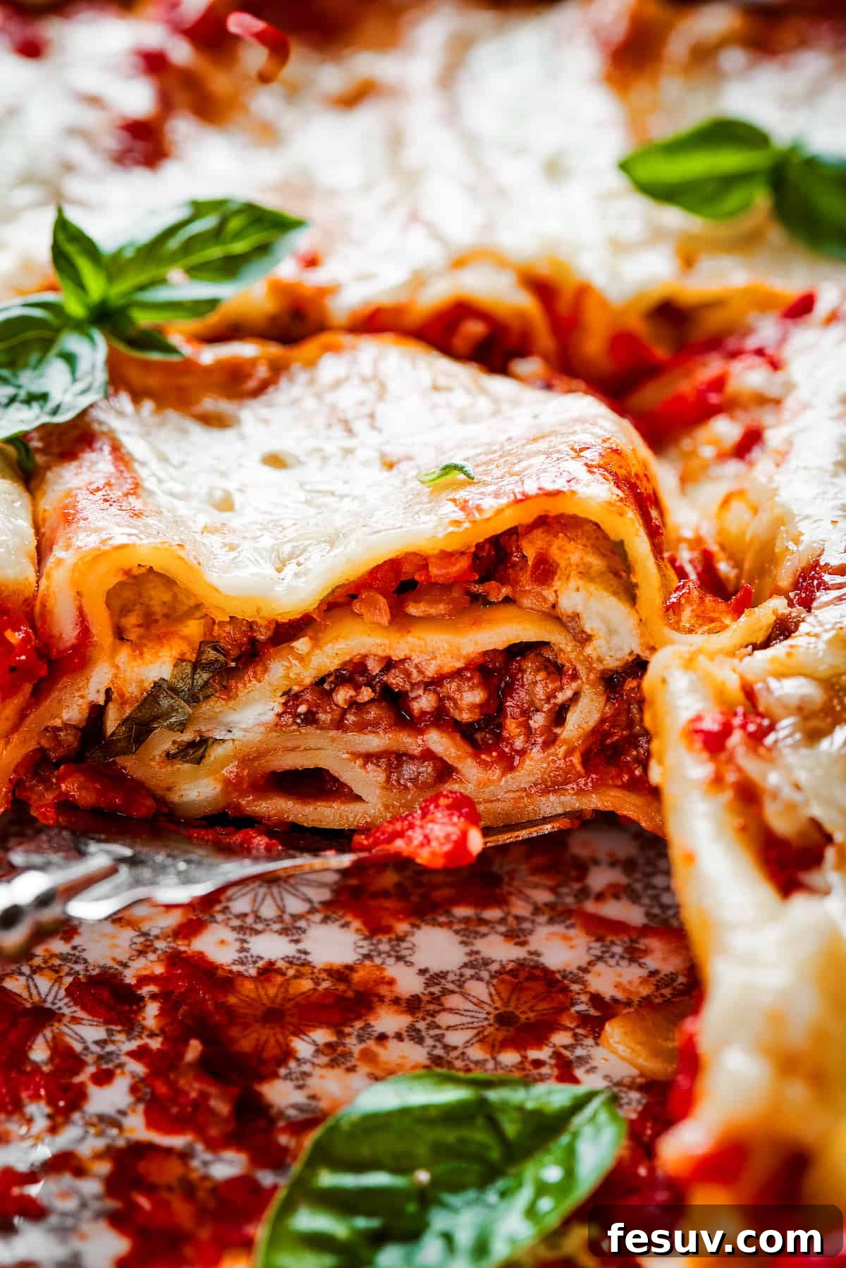 Easy lasagna roll ups have a beefy and herbaceous tomato sauce, a creamy cheese filling, tender pasta, and tons of melted cheese. Close up of a lasagna roll up on a plate topped with basil.
