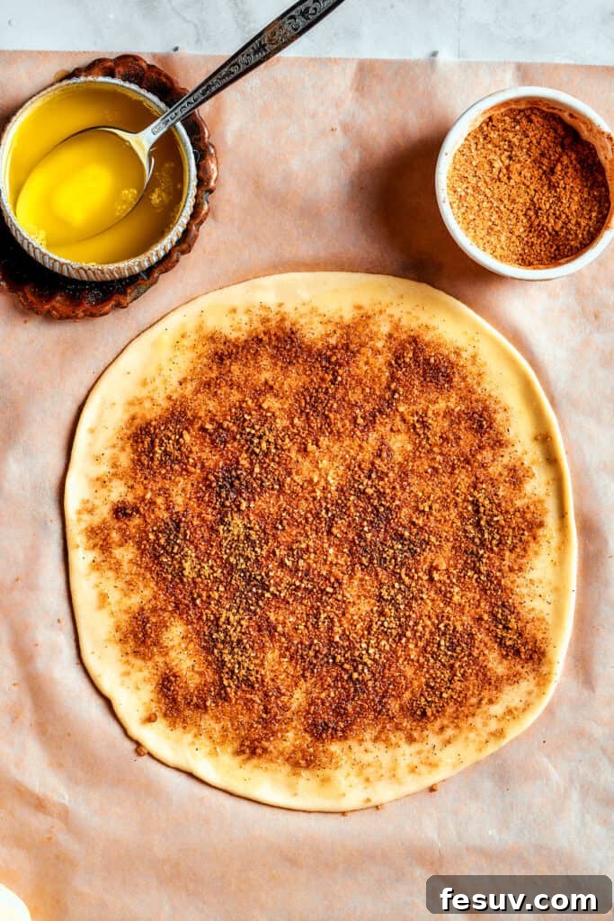 Overhead view of a perfectly rolled circle of Star Bread dough, lightly covered in cinnamon-sugar, with a bowl of melted butter and more cinnamon-sugar mixture nearby, illustrating the layering process.