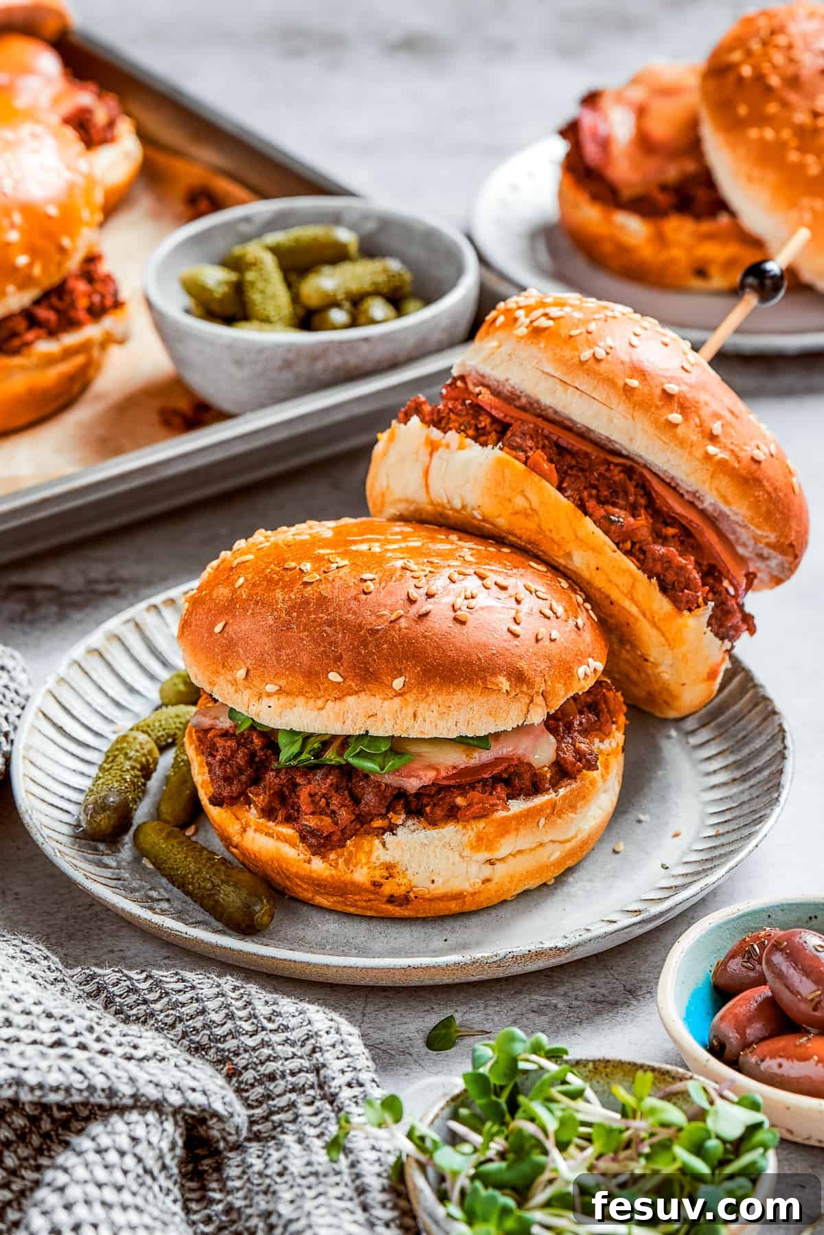 This easy Pizza Burger recipe is sure to become a family favorite! Meat filling is layered with pepperoni and melty mozzarella cheese. Two pizza burgers served on a plate with a few other burgers and pickles in the background.
