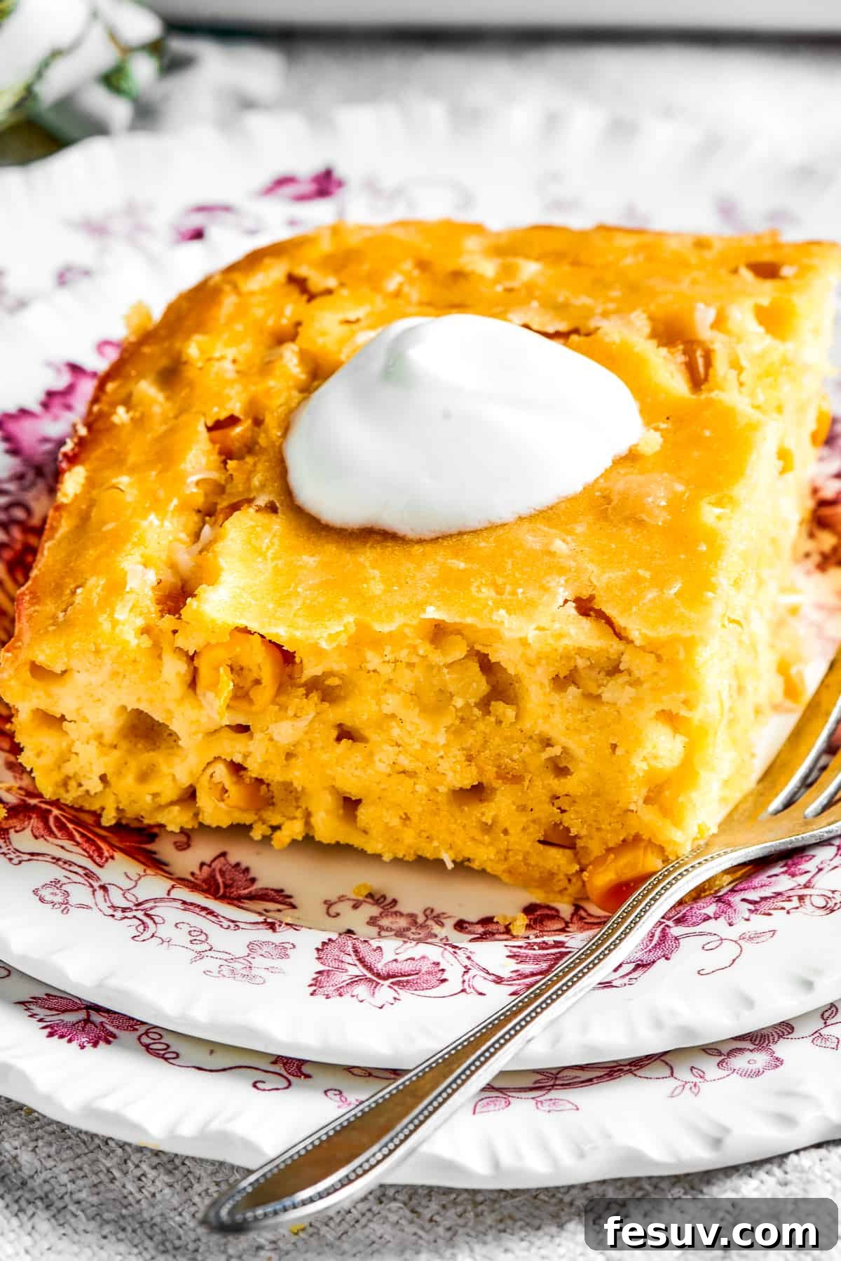 Golden Cloud Corn Souffle 10 A square of corn souffle served on a white dish, elegantly garnished with a dollop of sour cream and fresh herbs.