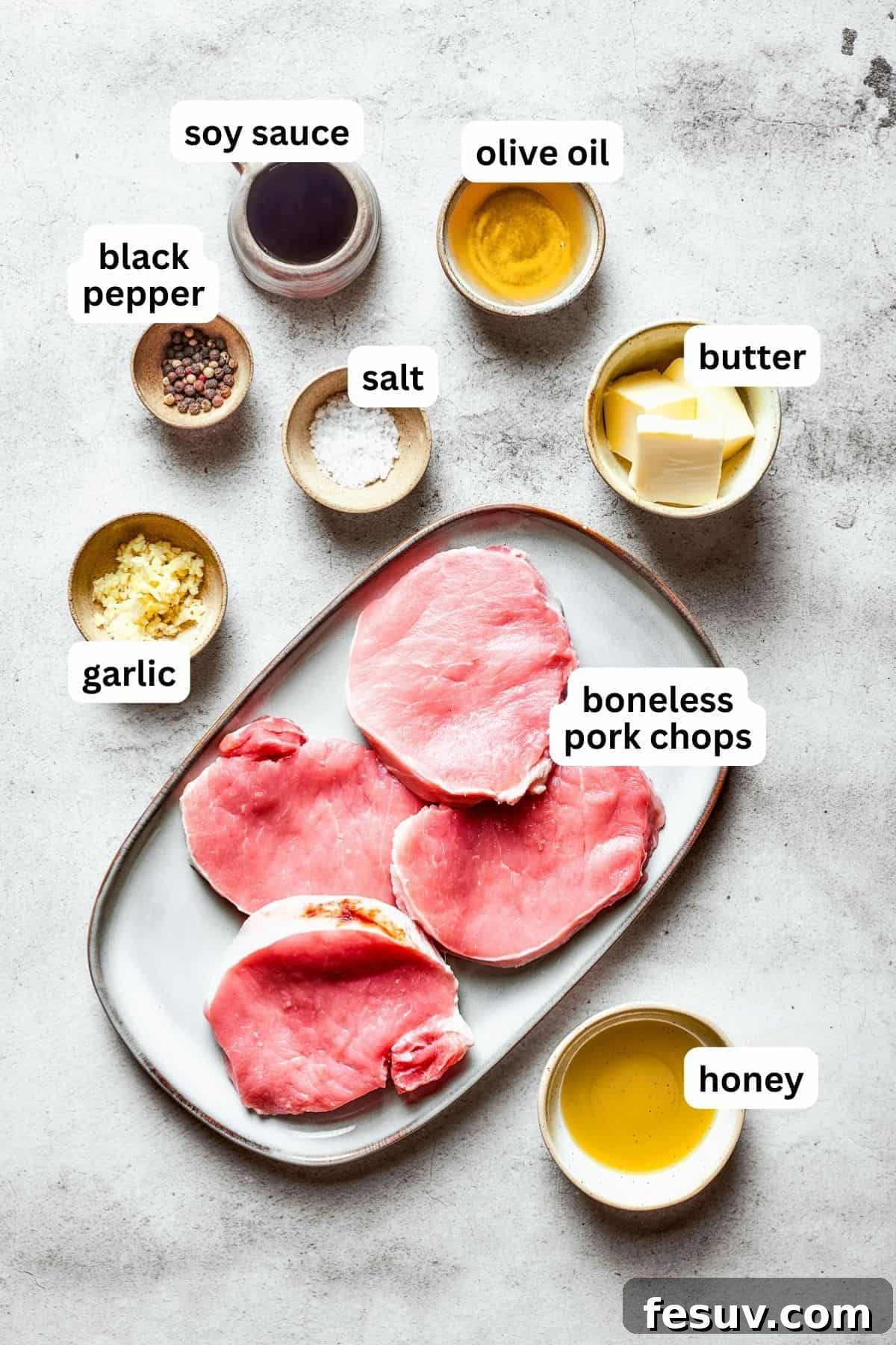 Overhead view of all the fresh ingredients used to make honey garlic pork chops, neatly arranged on a white surface, including pork chops, honey, soy sauce, garlic, butter, and parsley.