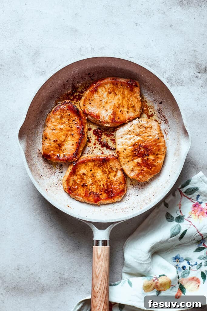Pork chops searing in a white skillet, acquiring a golden-brown crust.
