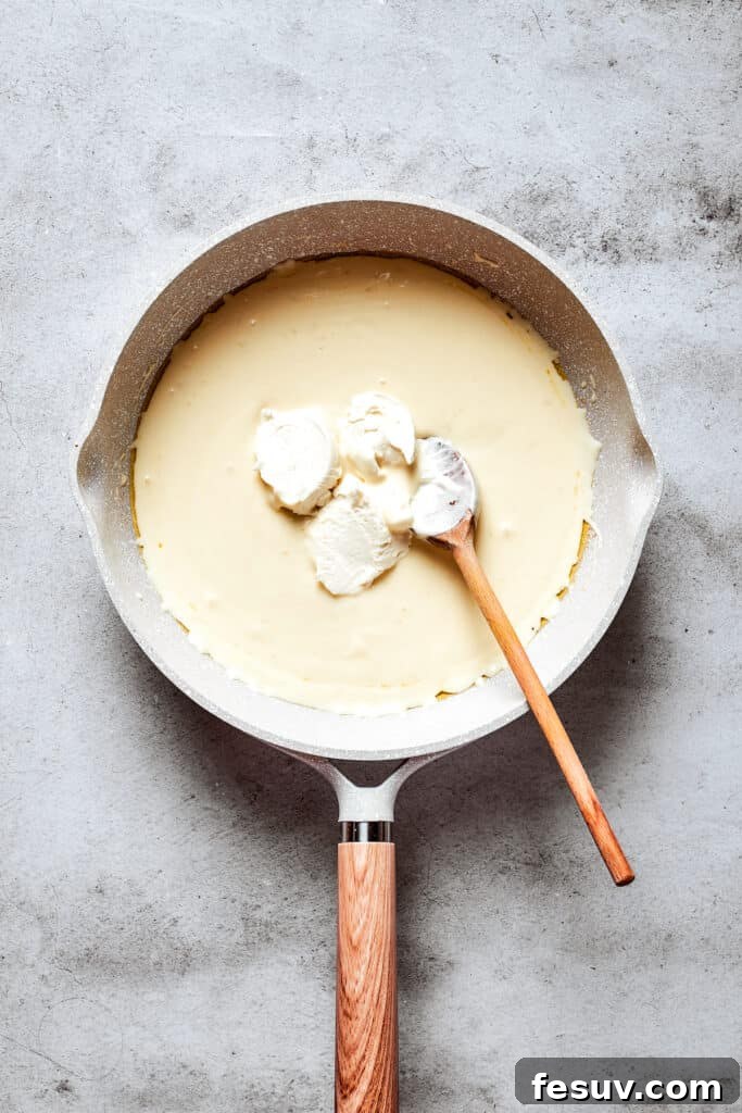 A wooden spoon stirs in cream cheese to a cream-butter mixture in a white skillet.