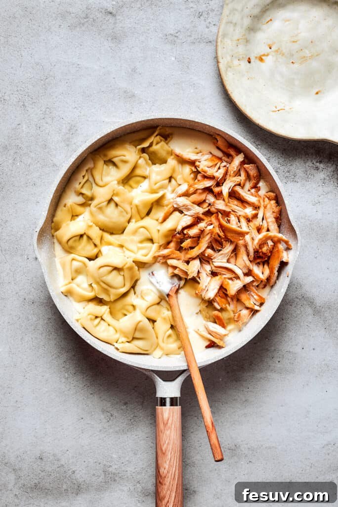 Cheese tortellini and shredded rotisserie chicken are stirred with a wooden spoon into a skillet of cream sauce.