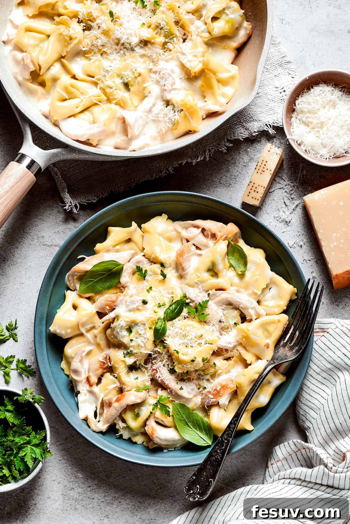 A blue bowl of chicken tortellini alfredo with a fork balanced on the edge of the bowl and a skillet of chicken tortellini placed near it.