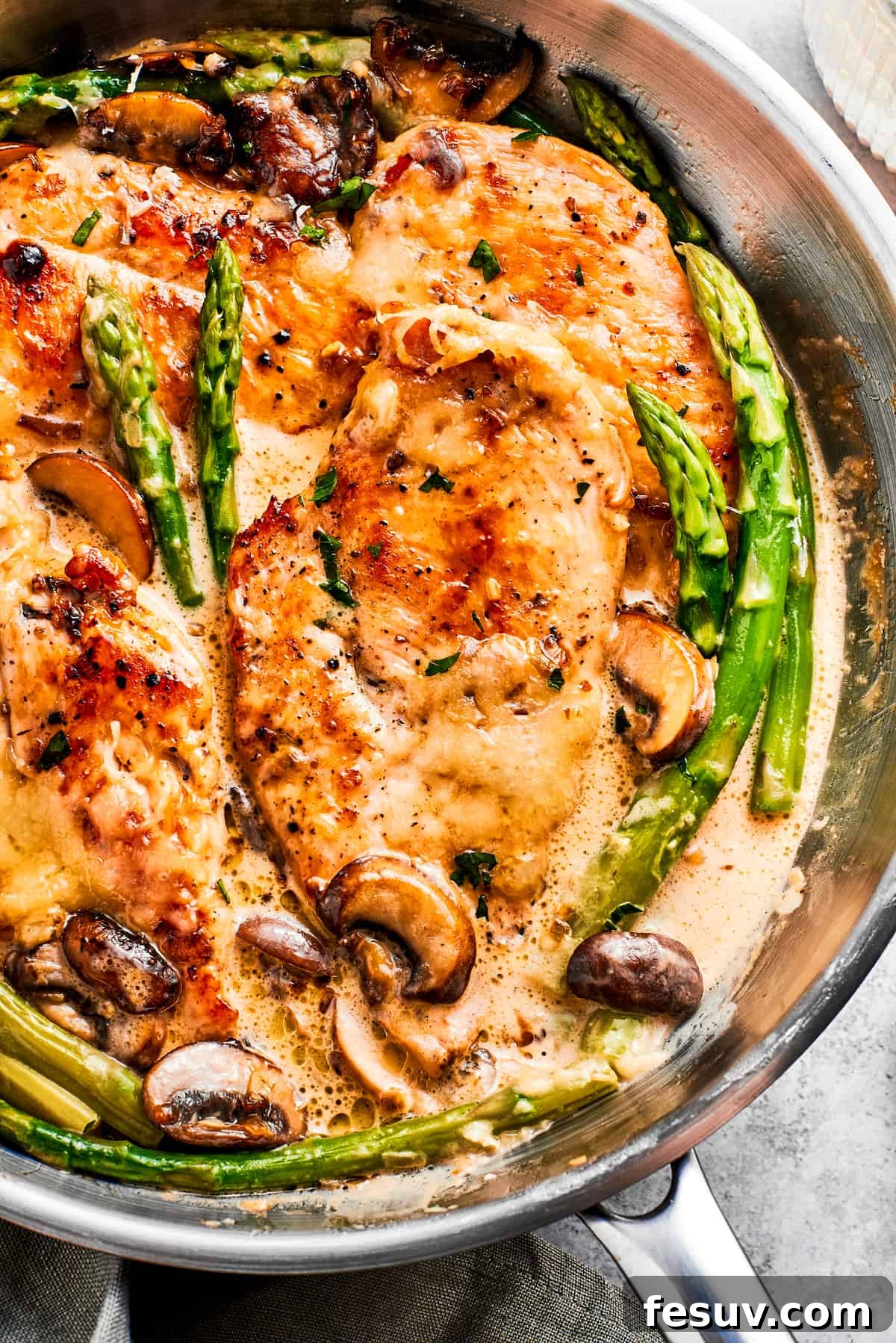 Homemade Chicken Madeira: A creamy, savory, and sweet dish with tender chicken, mushrooms, and melted mozzarella. Close-up photo of chicken Madeira and asparagus spears cooking in a skillet, showcasing the rich, creamy sauce and golden-brown chicken.
