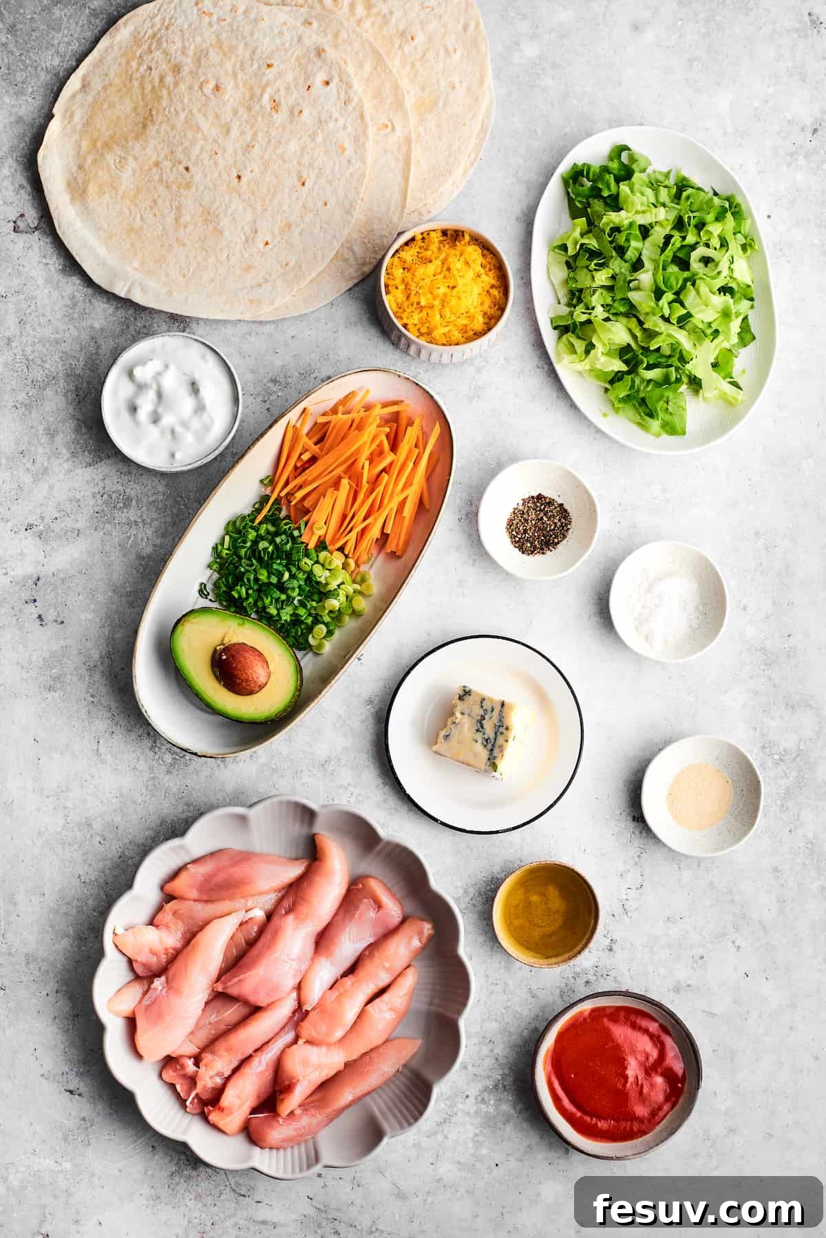 This Buffalo Chicken Wrap is packed with chicken tenders, cheese, lettuce, avocado, Ranch, and hot sauce in a warm flour tortilla. The ingredients for buffalo chicken wrap are shown portioned out: chicken tenders, olive oil, salt and pepper, garlic powder, hot sauce, ranch dressing, avocado green onions, carrots, tortillas, cheddar cheese, and blue cheese.