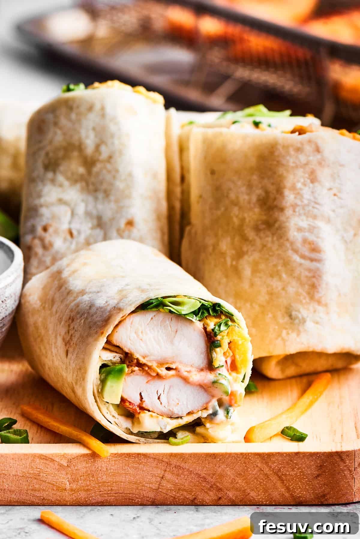 This Buffalo Chicken Wrap is packed with chicken tenders, cheese, lettuce, avocado, Ranch, and hot sauce in a warm flour tortilla. Sliced buffalo chicken wraps are displayed on a wooden cutting board with a bowl of ranch dressing nearby.