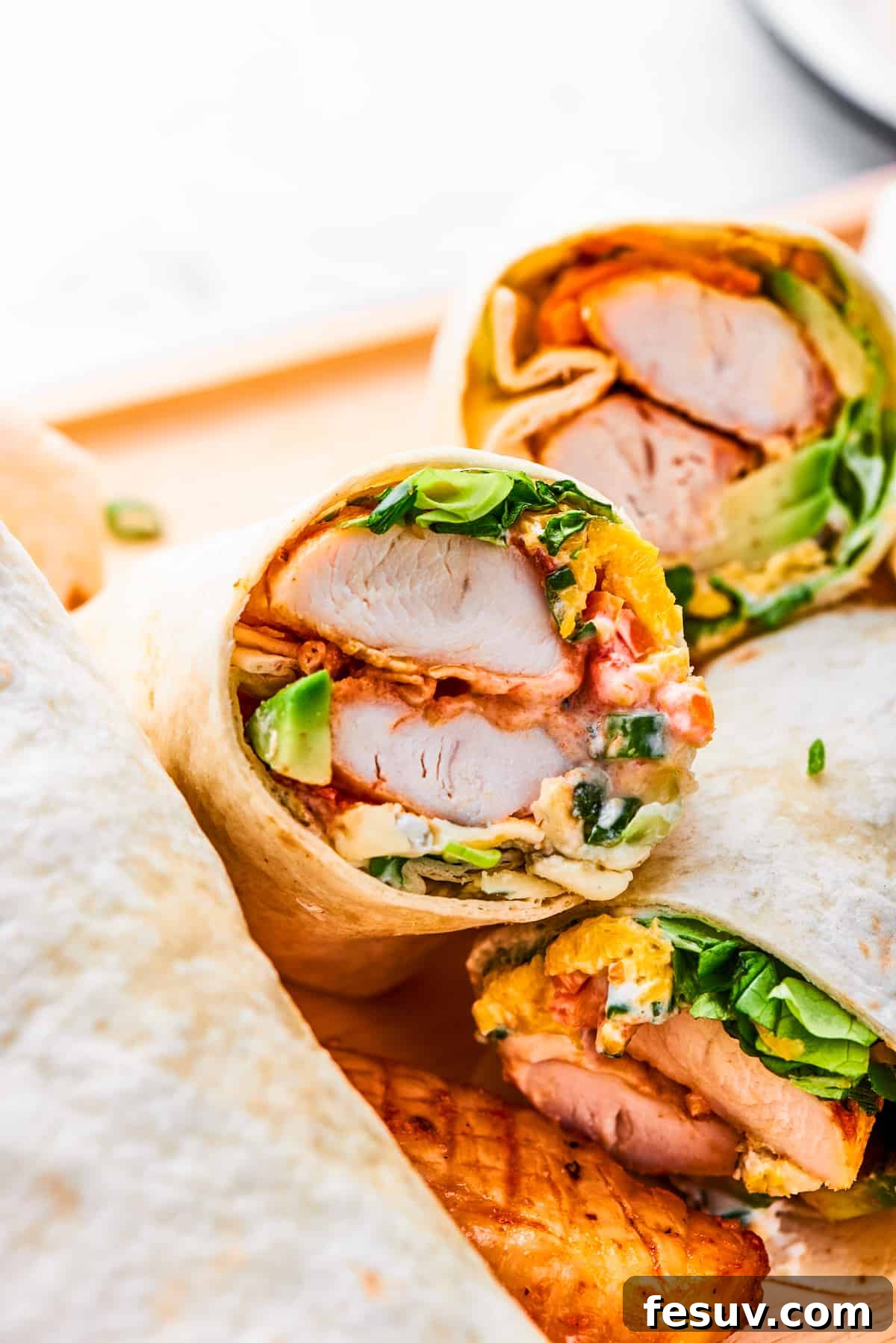 This Buffalo Chicken Wrap is packed with chicken tenders, cheese, lettuce, avocado, Ranch, and hot sauce in a warm flour tortilla. A halved buffalo chicken wrap shows the interior of chicken and vegetables.